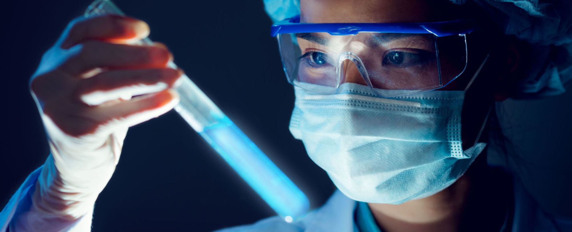 Harrington Discovery Institute Accelerates Breakthrough Science into Cancer Treatments and Cures. Harrington Advances Science to Medicine through Philanthropy. The image portrays a female scientist in a lab coat, mask and goggles looking at a science test-tube. .